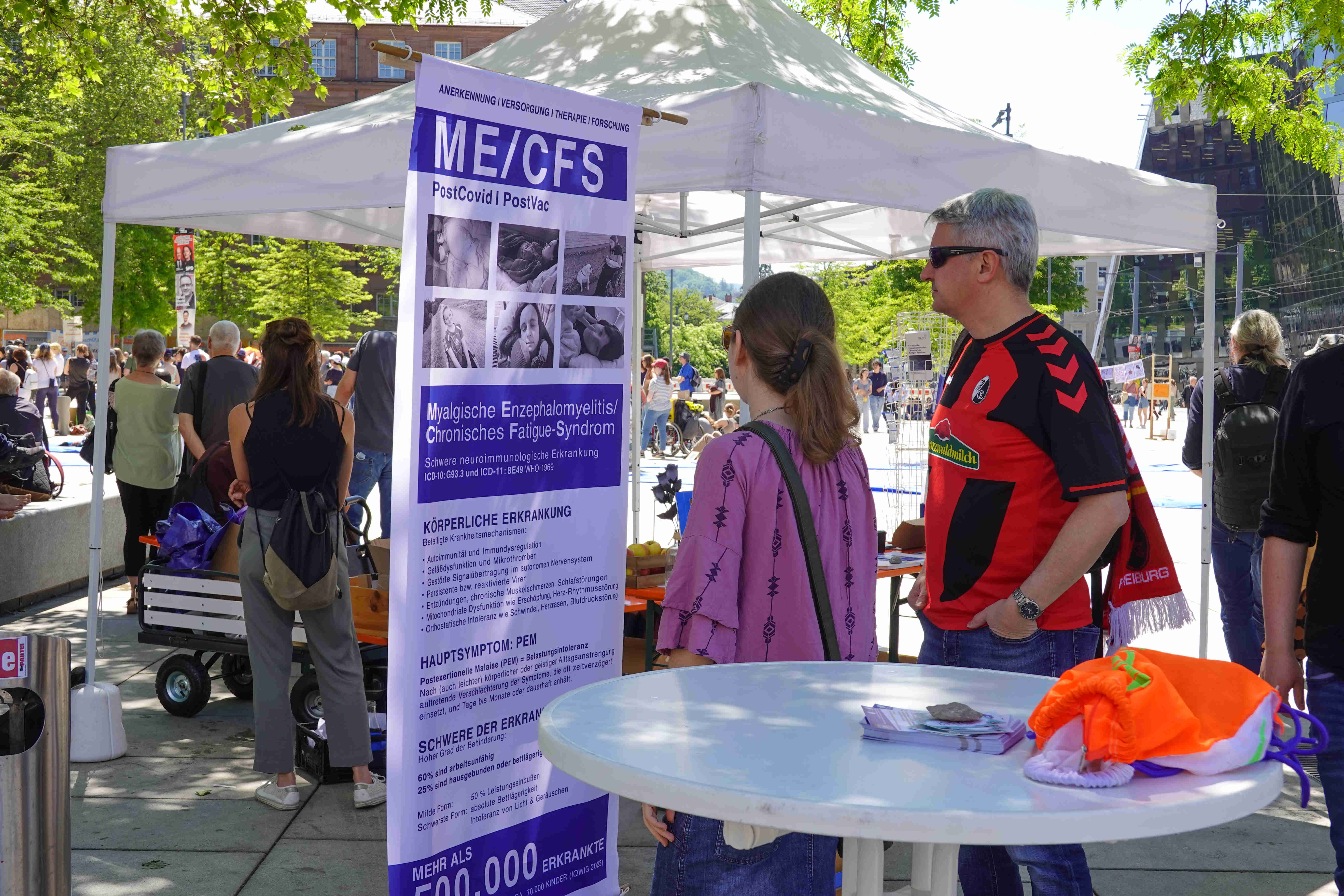 Bildbeschreibung: Foto des Infostands am Rand der Demonstration. Zwei Passant\*innen gucken sich ein Infobanner zu ME/CFS an. Das Banner zeigt sechs Fotos von Betroffenen und stichpunktartige Informationen über die Krankheit.